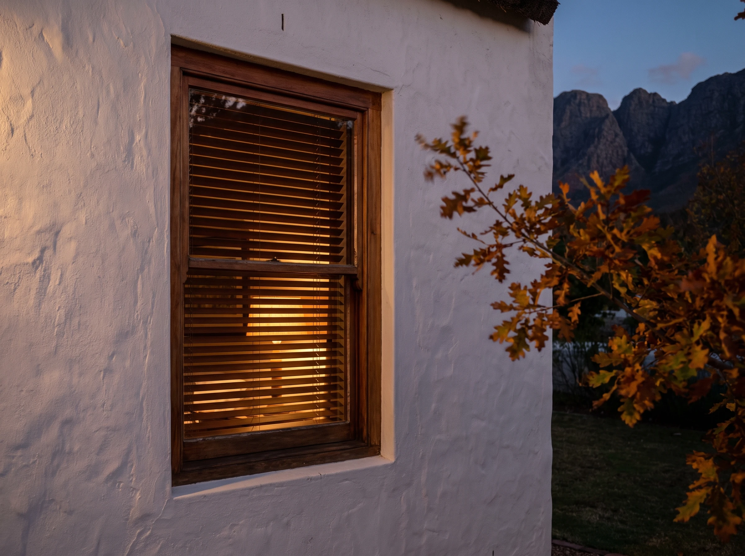 Deep-set sash window on a whitewashed protected Franschhoek village gable at dusk, a warm timber venetian blind glowing behind the glass, mountains fading to silhouette behind